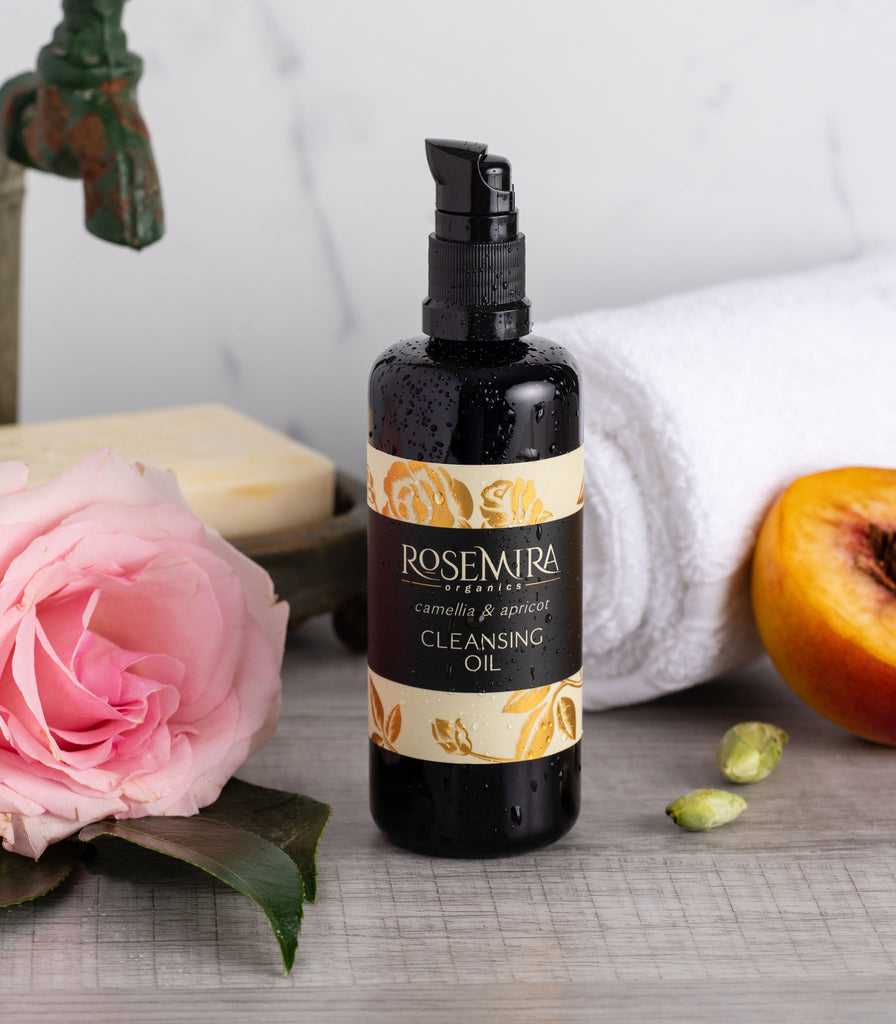 Water splashed bottle of cleansing oil with pink rose, apricot slice, and white towel.