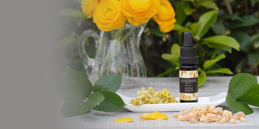Black bottle of vitamin c serum on white tray with ingredients and yellow flower bouquet.