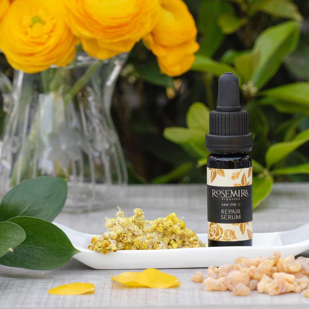Black bottle of vitamin c serum on white tray with ingredients and yellow flower bouquet.