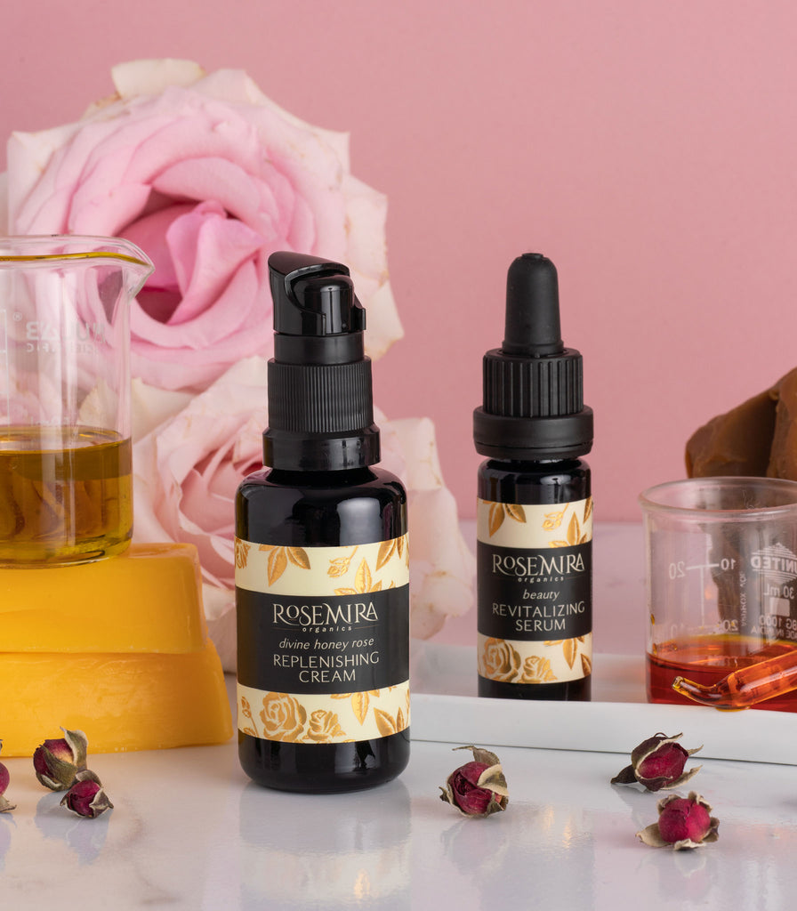 Organic face cream and serum bottles with ingredients, dried roses, on a reflective white surface.