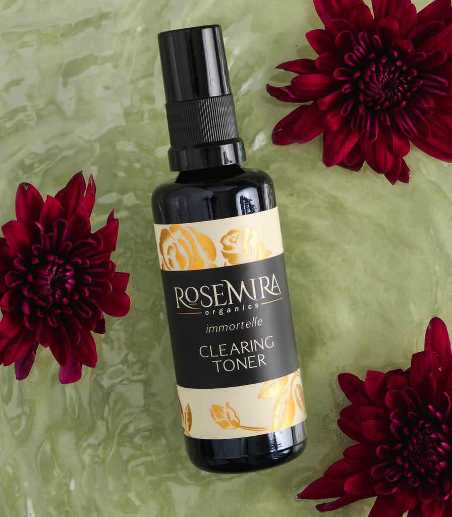 Immortelle Clearing Toner with red dahlias on a wet green surface.