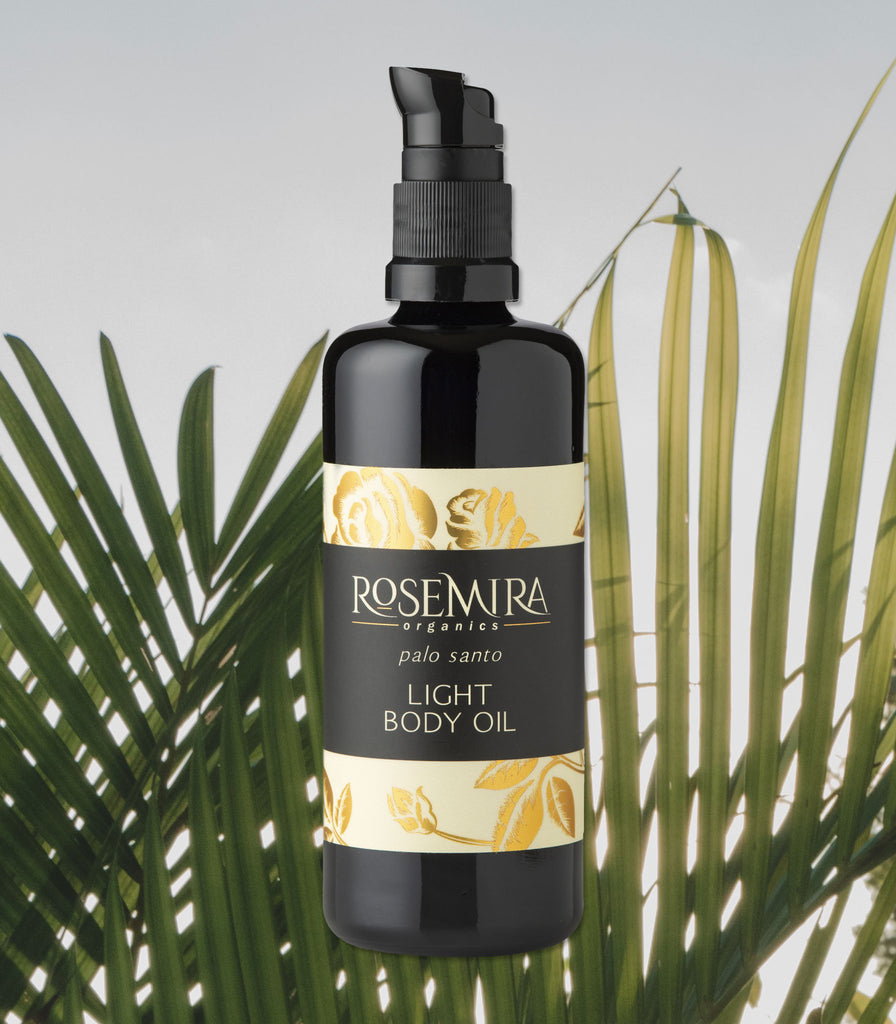 Palo Santo Body Oil in glass bottle floating over sun-tinged palm leaves.